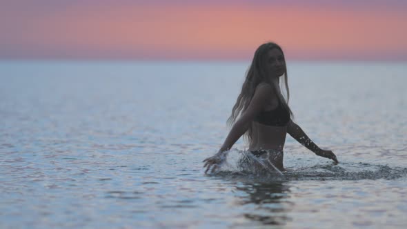 A Girl with Blond Hair in a Black Swimsuit Splashes to the Sides While Sitting in an Estuary on a alt