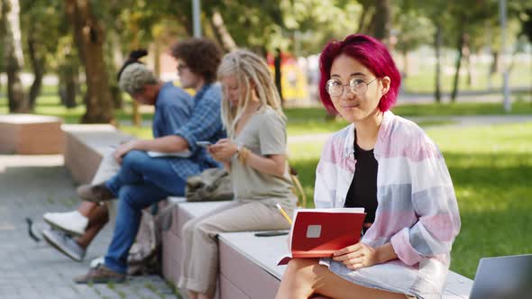 Young Asian Female Student Holding Notepad and Smiling at Camera Outdoors alt