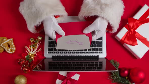 Top View Santa Hands in White Gloves are Typing on Keyboard Laptop By Red New Year Decorated Table alt