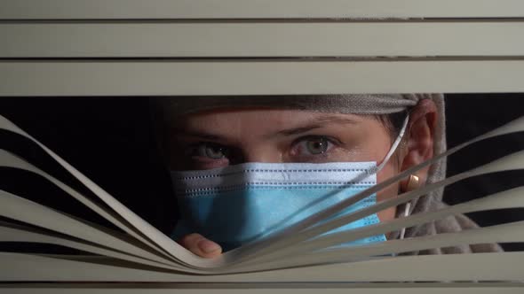 Woman in protective mask looking out the window through the blinds to street alt