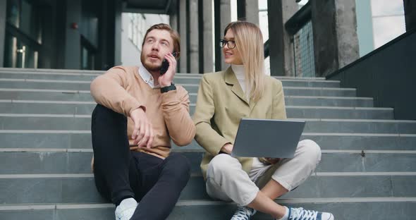 Man and Woman Sitting on Stairs in Urban City Center Working on Laptop, Talking on Mobile phone alt