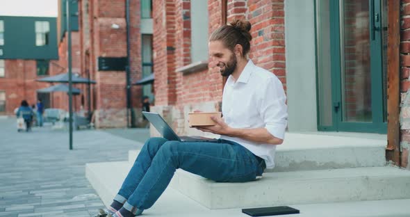 Handsome Bearded Businessman Working on Laptop and Eating Salad at the Office Center Outdoor. alt