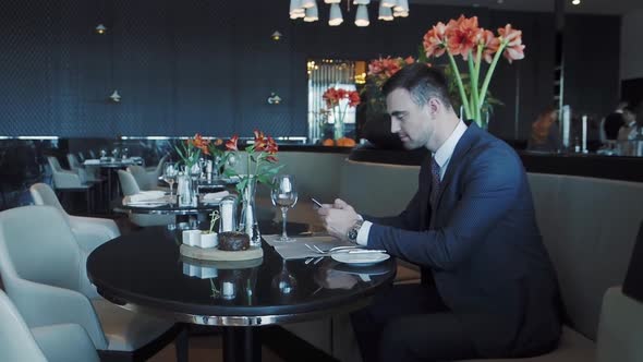 Businessman Sits at a Table in a Restaurant and Uses His Smartphone