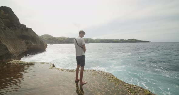 Back View of a White Male Tourist Standing on the Flooded Cliff Beach and Looking Into an Ocean at alt