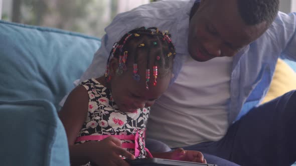 African American father and daughter playing in living room, Kid girl and dad playing on a tablet alt