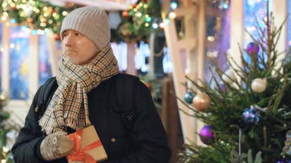 An Elderly Man with a Gift in His Hands Stands at a Christmas Market Waiting for a Date on a Snowy