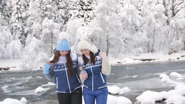 Two Lovers Girls Holding Each Other's Hands are Walking Near the River in a Snowy Forest alt