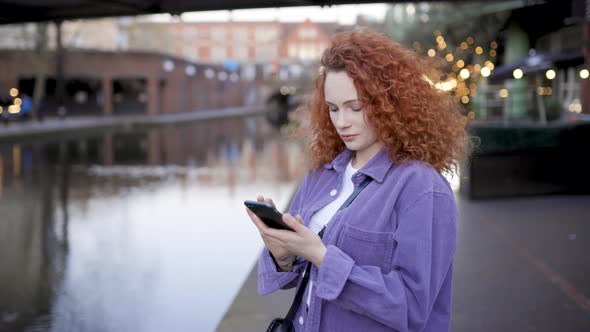 Woman with curly hair standing by river using smartphone