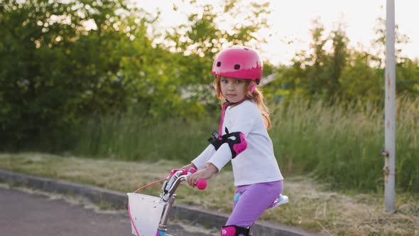 Cute Light Hair Little Girl in Pink Helmet in Elbow and Knee Pads Sits on Bicycle and Starts to Ride