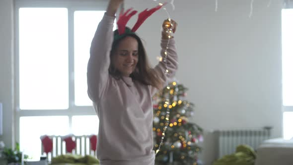 Young Beautiful Woman Wearing Reindeer Antlers Headband and Garland Dancing Next to Christmas Tree