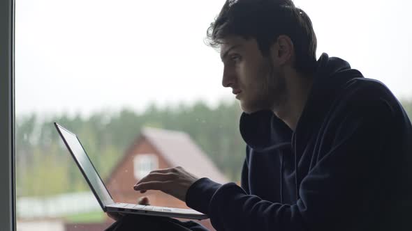 A Young Man is Typing on a Laptop Sitting at the Window Against the Backdrop of a Picturesque Rural alt