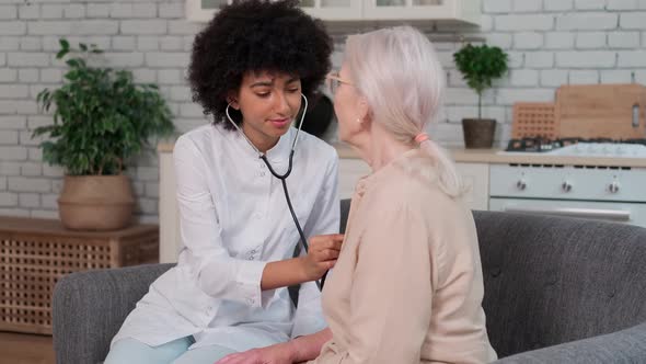 Afro American Woman Doctor Listens to Breath of Senior Woman Using Stethoscope While Sitting on Sofa alt