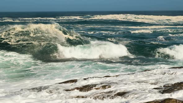 Wave collides dramatically on rocky shore, big splash; low angle static alt