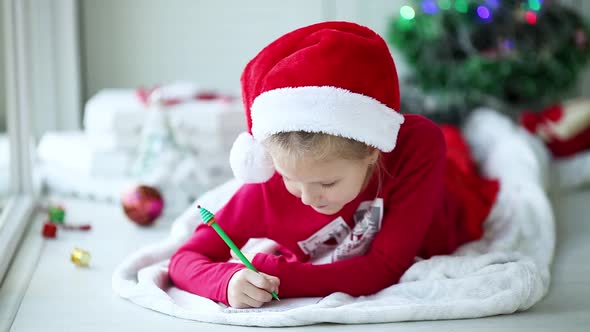 A smiling girl in a Santa hat writes a letter, dreams of gifts to Santa Claus.
