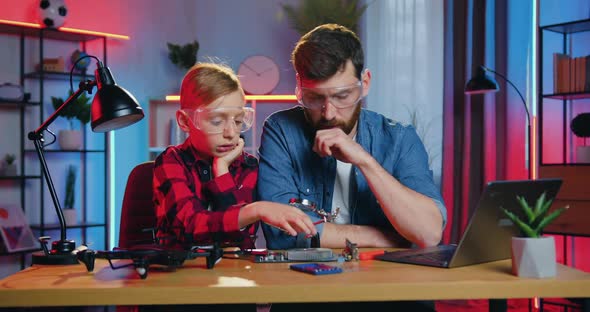 Bearded Man and His Teen Son Both in Glasses Talking while Trying to Repair Electronic Panel alt