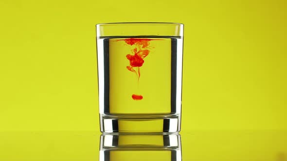 Two Red Drops of Ink Dripping Into Clear Water in Glass Standing on Yellow Background in Slowmotion alt