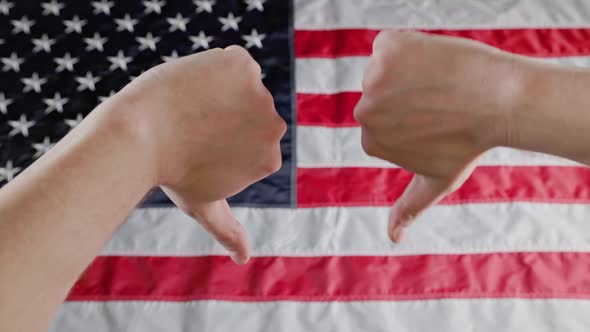 Thumbs Up and Thumbs Down Gestures Made with Two Caucasian Hands in Front of Blurry US Flag alt