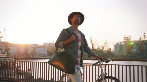 A Young Stylish Hipster with Hat and Glasses Walking with His Bicycle on Sea Port Background During alt