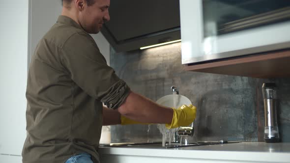 Young Man in Gloves Washing Dishes While Standing in Kitchen. alt