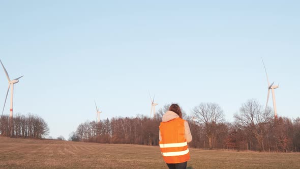 Female Engineer in Orange Vesta Goes to Wind Turbines with a Tablet to Checks Their Operation alt
