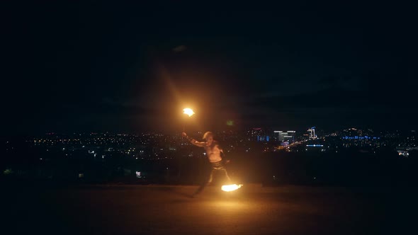 Young Blond Male Dances with Two Burning Torches in the Middle of the Night with City Skyline in alt