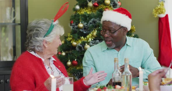 Happy diverse senior couple in santa hats talking at christmas dinner table at home alt