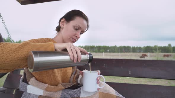The girl pours tea into a white mug from a thermos while sitting on a swinging bench.