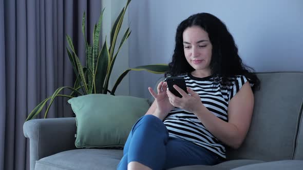 Woman Typing on Phone Sitting on Sofa at Home, Businesswoman Sits at Home Types on Smartphone Checks alt
