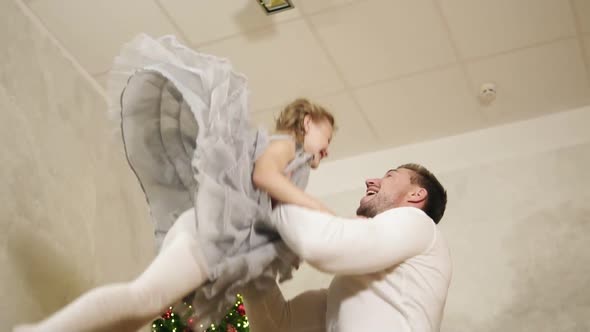 Young Father is Throwing His Adorable Daughter in the Air at Home with Decorated Christmas Tree on alt