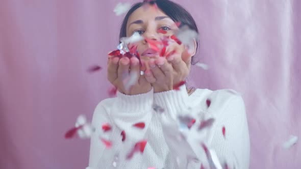 Smiling Funny Young Brunette in a White Sweater Sends Air Kisses Blowing on Red Hearts on a Pink alt