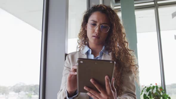 Biracial businesswoman sitting by desk and using tablet in modern interiors alt