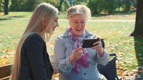 Beautiful Blonde Girl with Elder Woman Sitting on Bench and Looking Something on Smartphone alt