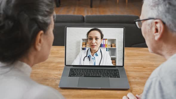 Back View of Aged Couple Looking at Screen of Laptop and Talking to Doctor Woman Using Online Video alt