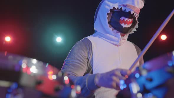 A Male Musician Disguised As a Shark Plays a Drum Kit in a Dark Studio with Bright Colored Lights alt