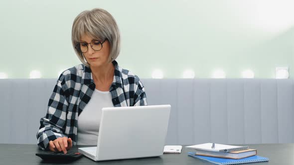 An Elderly Beautiful Woman Works at Home at a Laptop in the Home Office