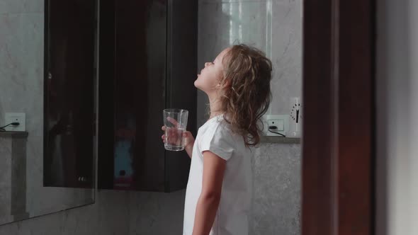 Little curly child girl rinses her mouth with water in bathroom, side view.