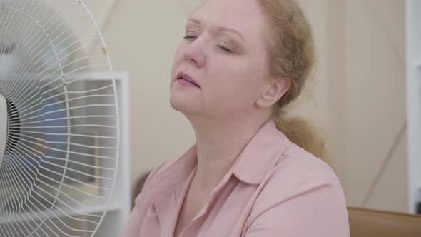 Close-up of Senior Woman Using Electric Fan Indoors. Portrait of Ill Caucasian Lady Having Fever or alt