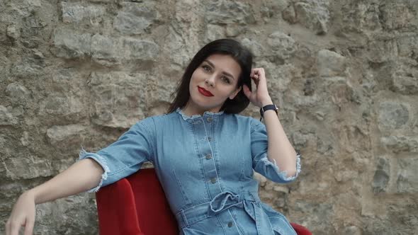 Beautiful young smiling brunette woman with red lipstick sits on an armchair alt