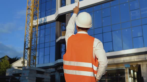 An Engineerarchitect in a White Shirt Helmet and Orange Work Vest Stands with His Back to the Camera alt