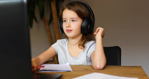 Cute little girl with headphones using laptop to study at home alt
