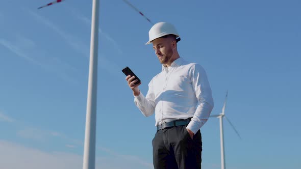 An Engineer in Business Clothes and Helmet Works in a Cell Phone Against the Background of Wind alt