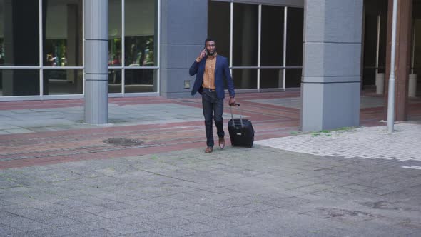 African american businessman using smartphone walking with suitcase in street alt