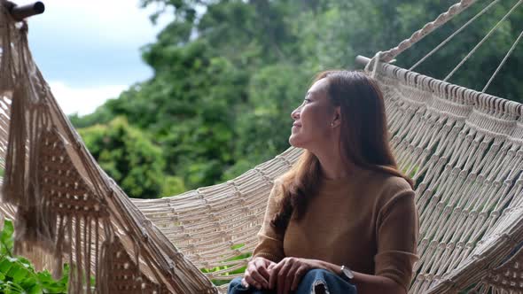 A young asian woman sitting and relaxing on hammock while looking at a beautiful nature view