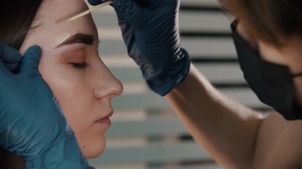 Brow Master in Black Mask Applying Wax on the Brow of Young Woman Model alt