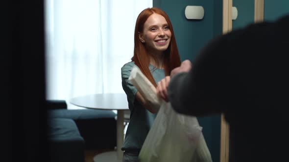 Charming Redhead Young Woman Gratefully Receiving Bag with Healthy Food at Home From Delivery Man alt
