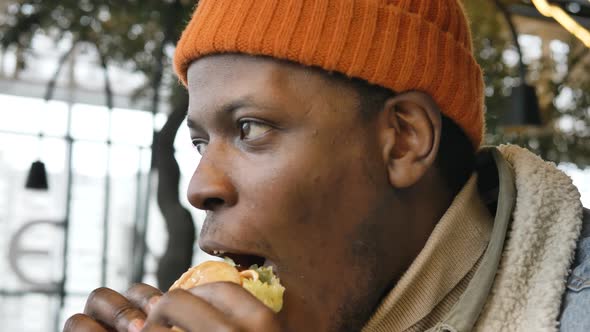 Black Guy in Jacket and Hat Tastes Delicious Burger in Cafe alt
