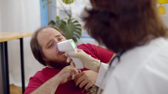 Doctor in Protective Mask Measures Temperature with Noncontact Thermometer To Patient at Home alt