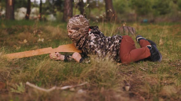 Young Boy in Camouflage and in Balaclava with Wooden Gun Lays on the Grass