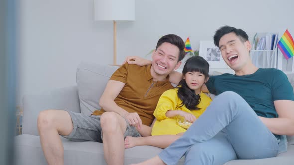 Asian male gay family with young daughter sit on sofa and watch movie in living room together. alt