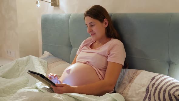 Portrait of Young Pregnant Woman with Big Belly Lying in Bed with Tablet Computer and Browsing alt